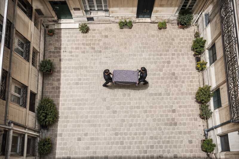 Deux déménageurs traversent la cour intérieure pavée d’un immeuble